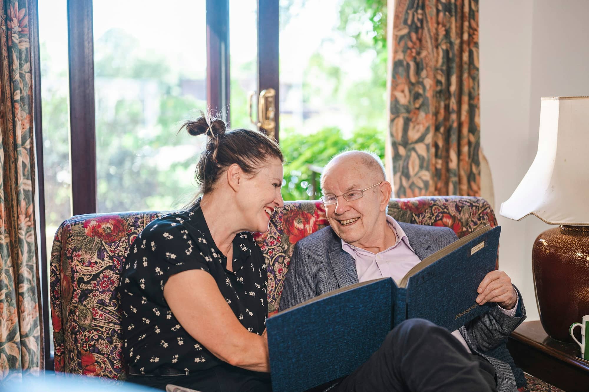 A woman and an older man laughing together over a photo album at home